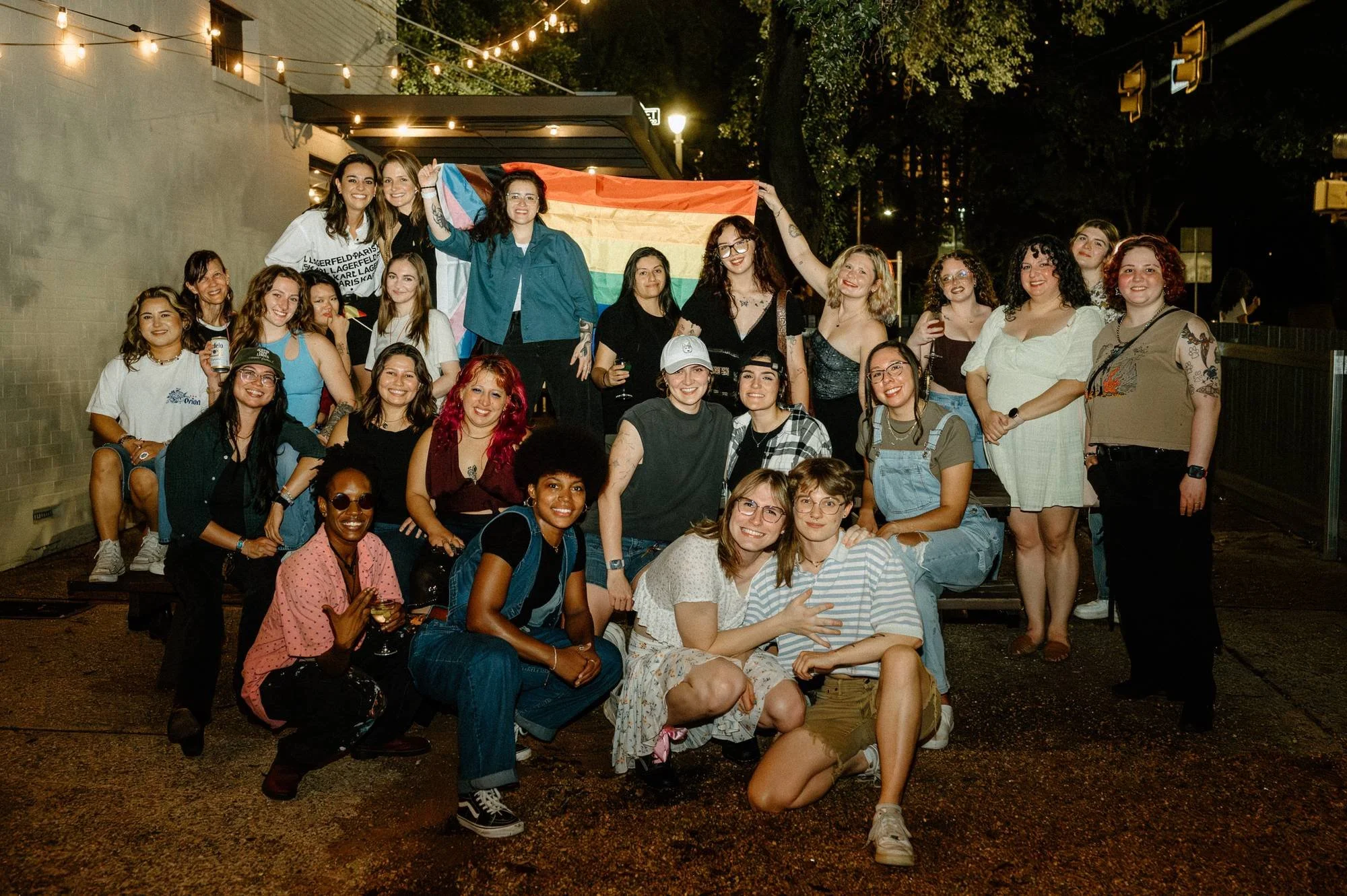 A group of diverse women gathered outdoor at night, smiling for a photo. Some are holding drinks, and one woman is holding a rainbow flag. There are string lights above and trees in the background.