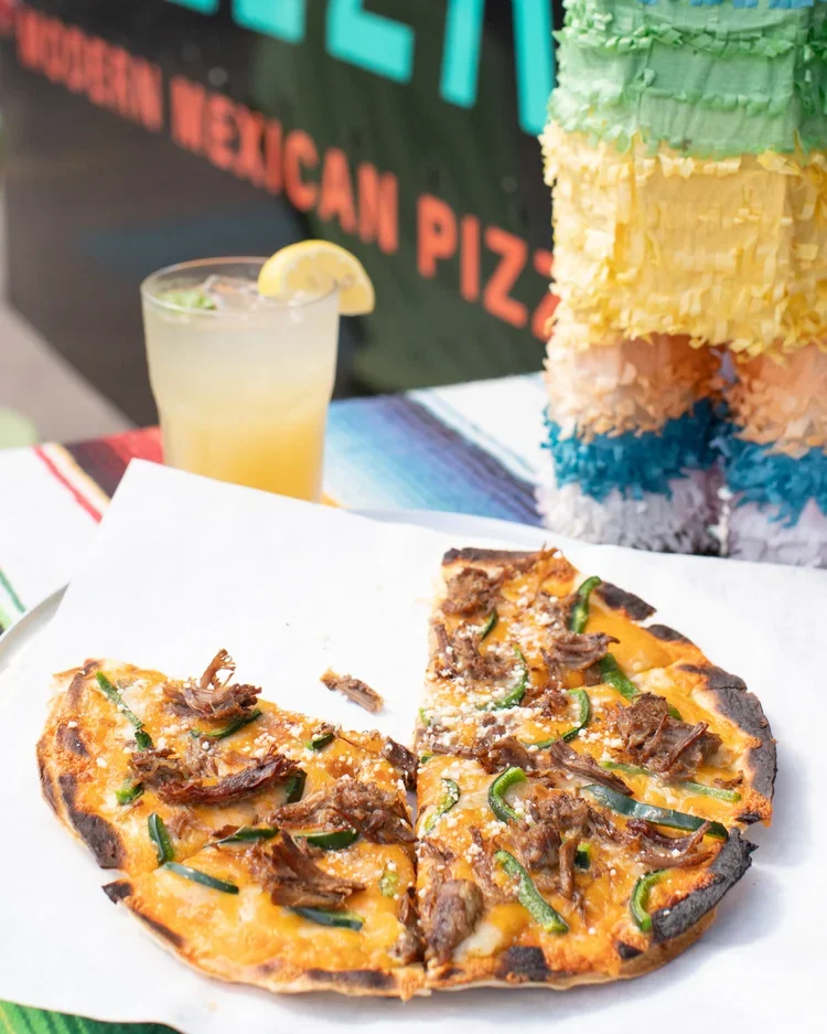 A sliced beef and jalapeño pizza on a white paper with a lemon drink and colorful Mexican decorations in the background.
