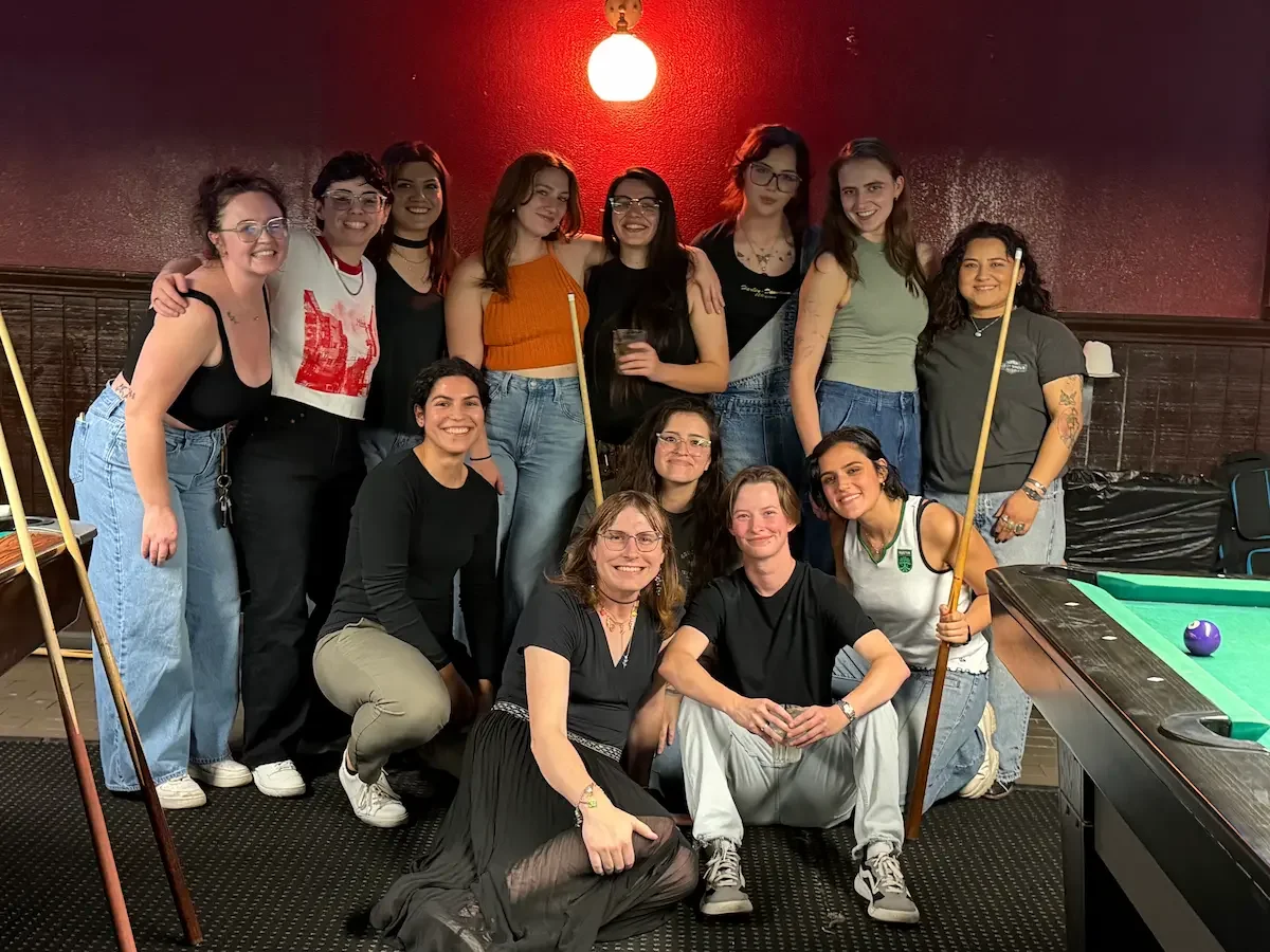 A group of 13 women posing together in a bar or game room, some holding pool cues, with a pool table to the right, and a red wall with a hanging light in the background.