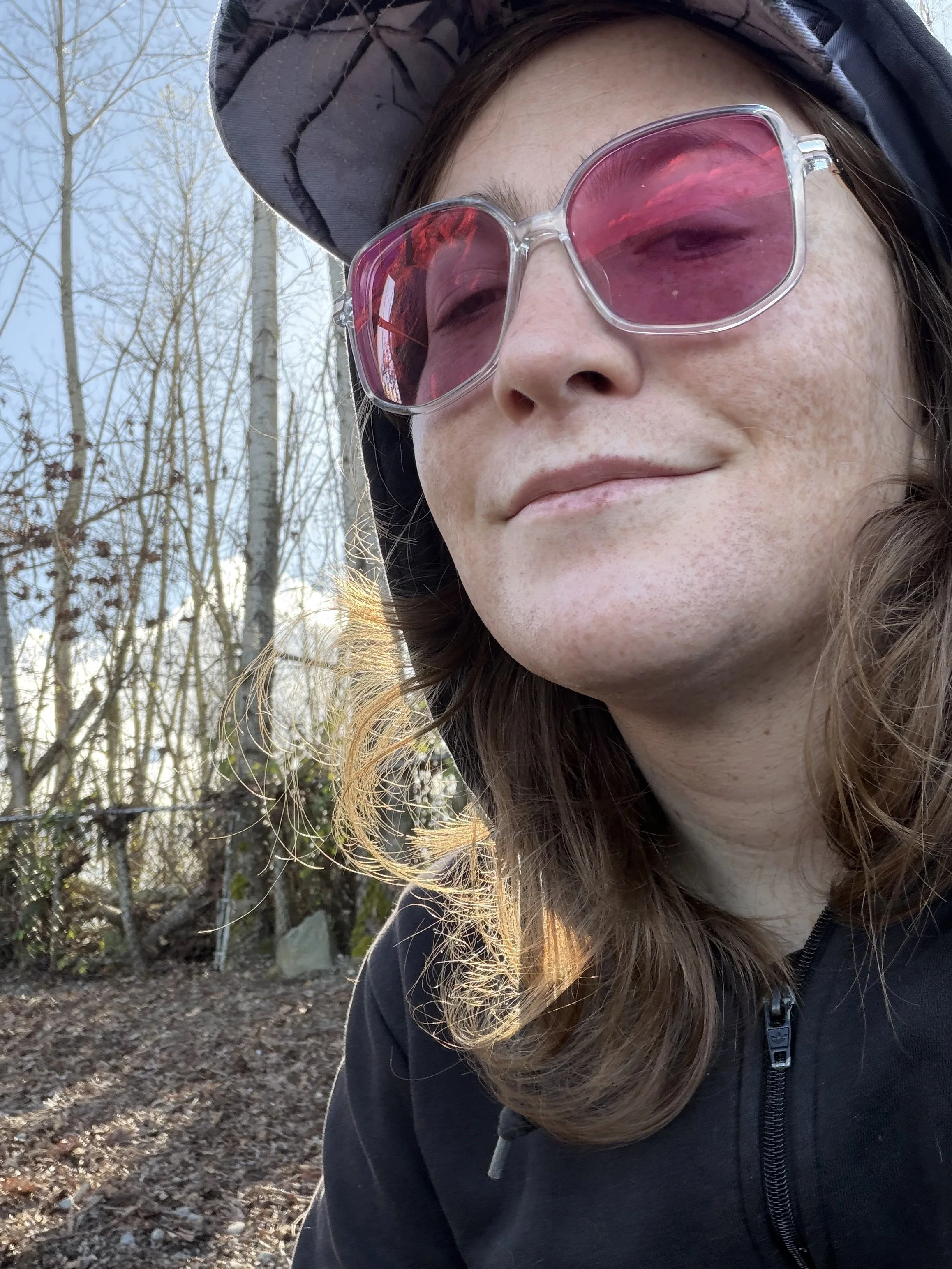 A woman outdoors wearing pink-tinted sunglasses and a wide-brimmed hat, with trees and a dirt ground in the background.