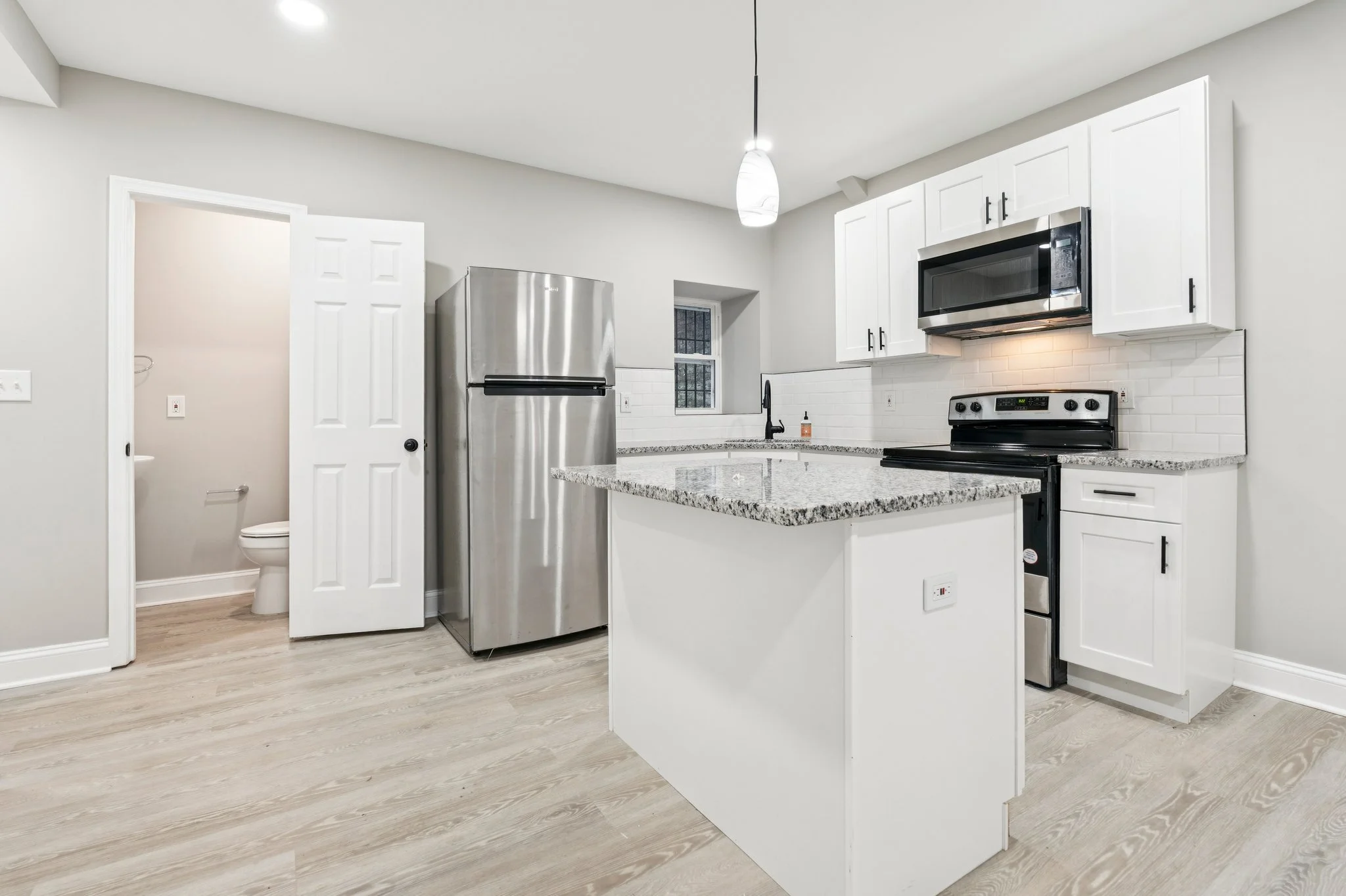 Modern kitchen with white cabinets, granite countertops, stainless steel appliances, a small window, and a visible bathroom with a toilet.