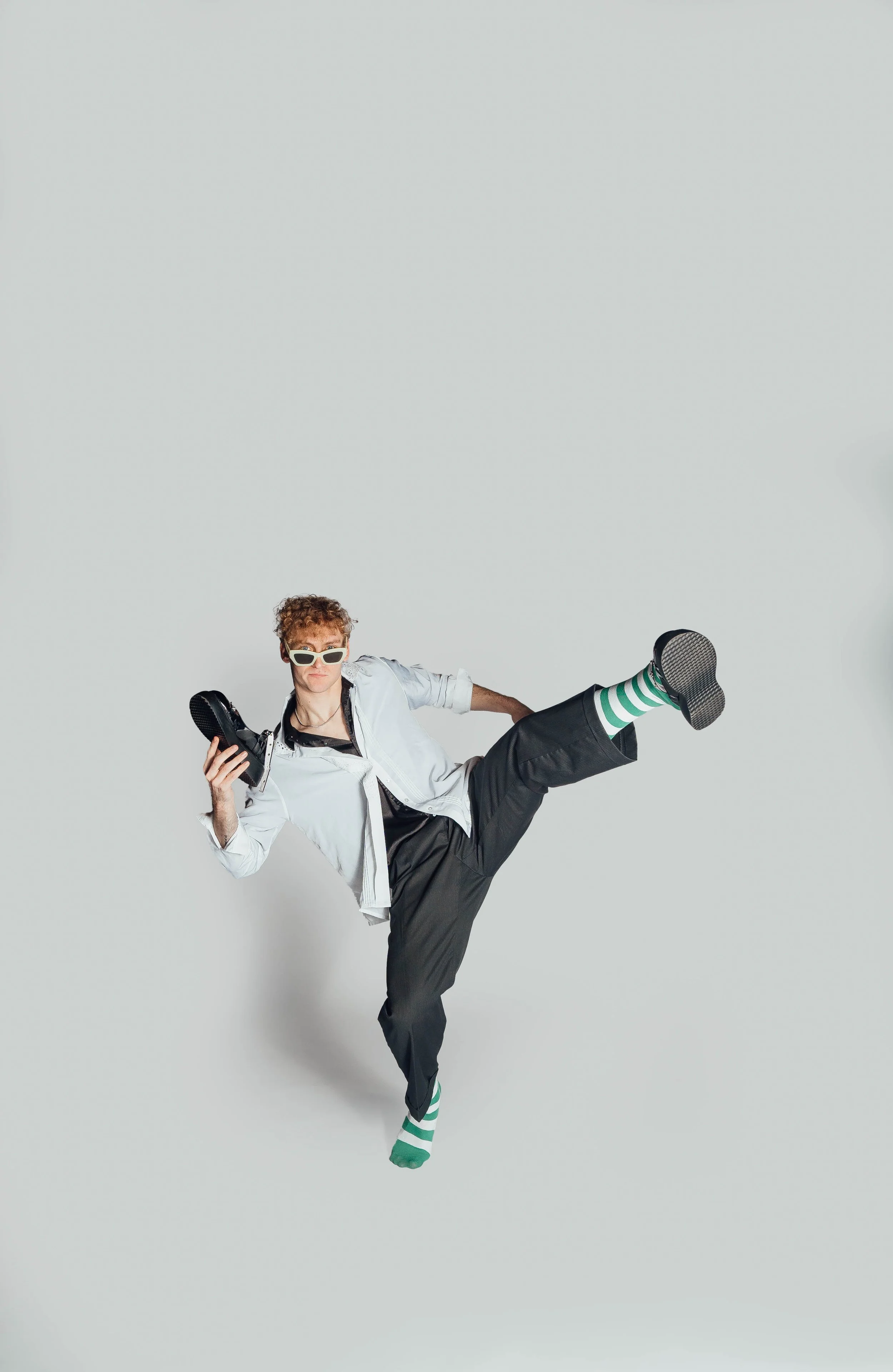 Young man with curly hair, sunglasses, white shirt, black pants, and green-and-white striped socks holding a black shoe in one hand, standing against a plain light gray background.