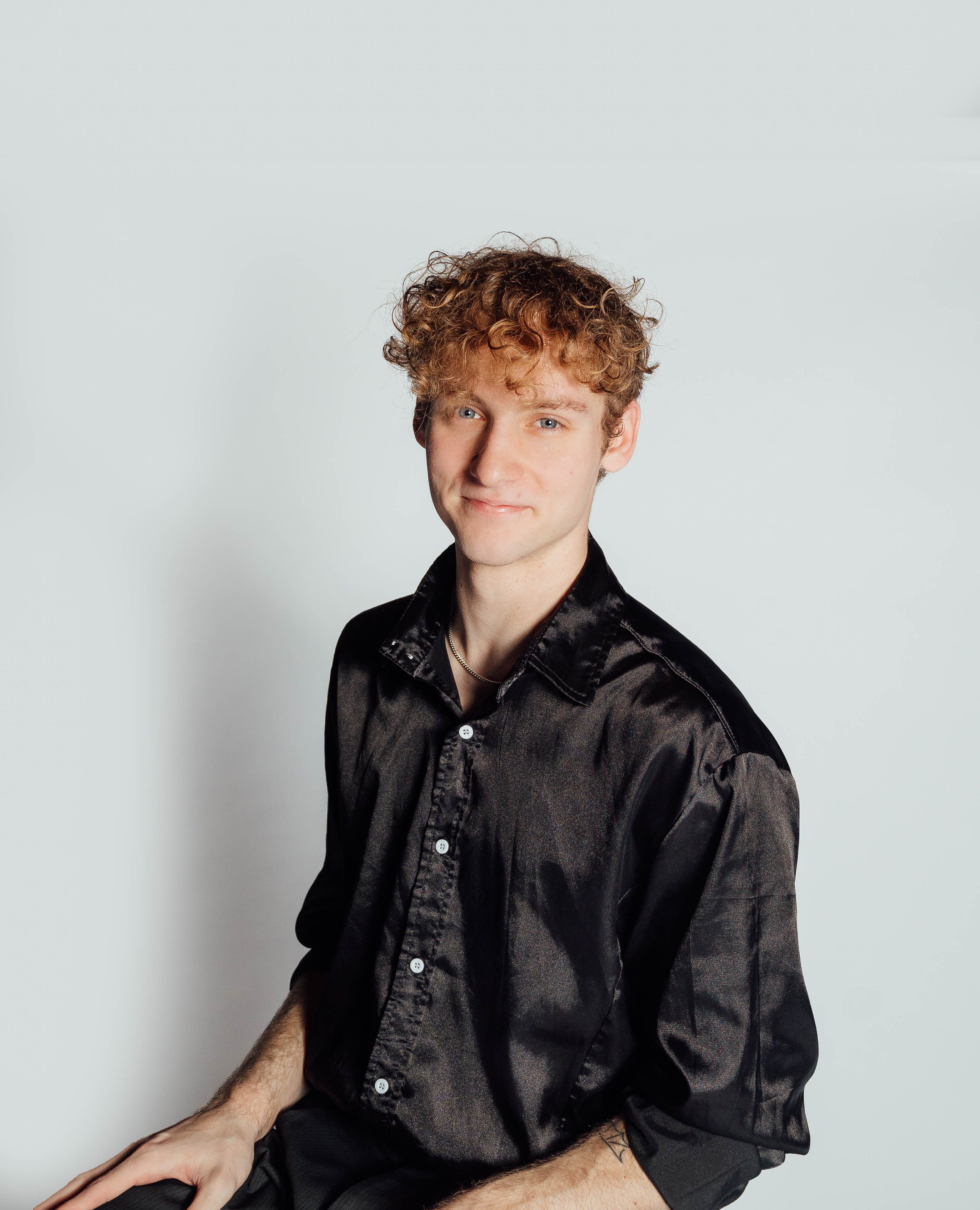 A young man with curly blonde hair, blue eyes, and fair skin, wearing a black satin shirt, smiling softly, sitting against a plain white background.