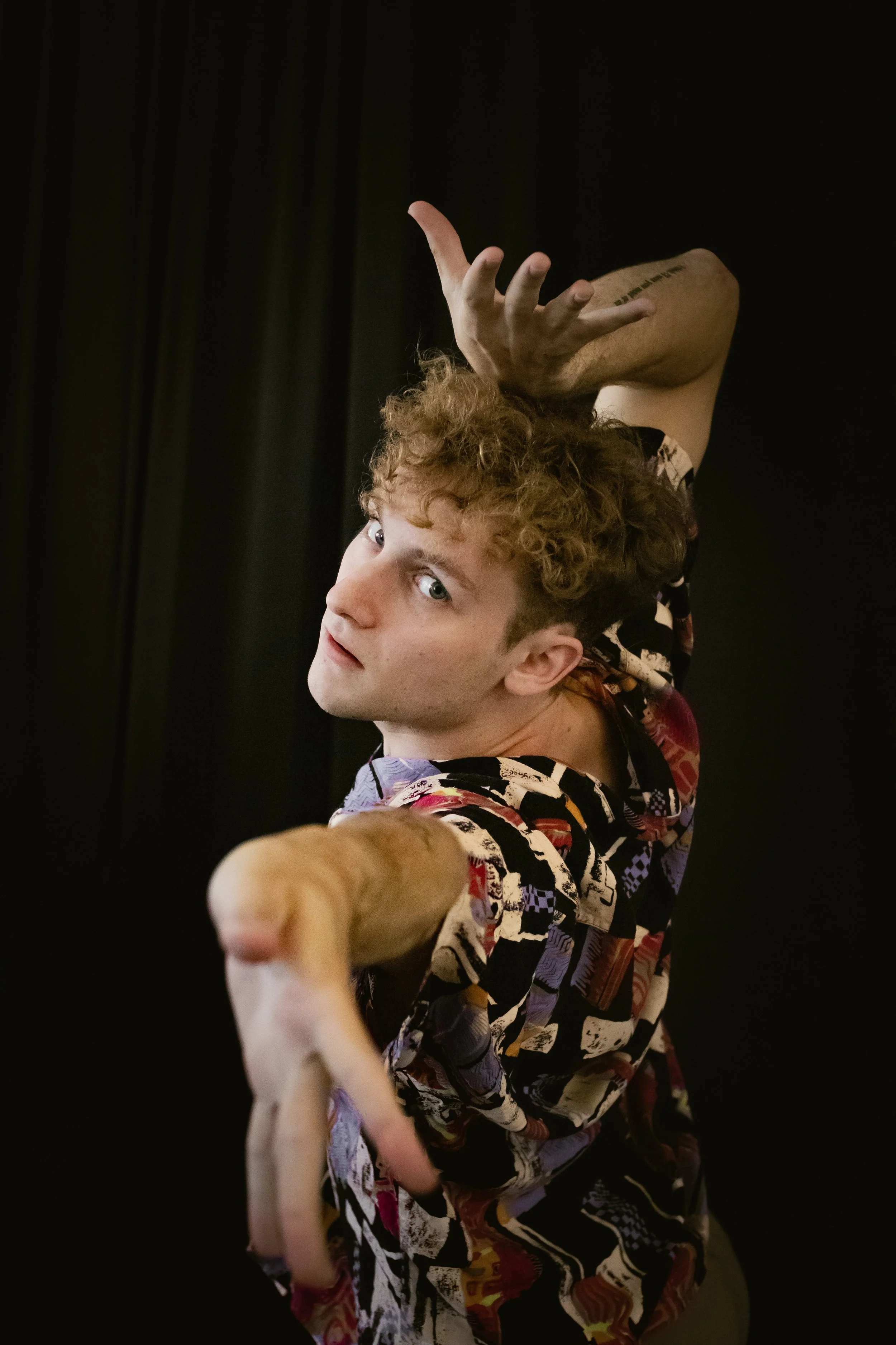 Young man with curly hair in a colorful shirt, reaching towards the camera, set against a black background.