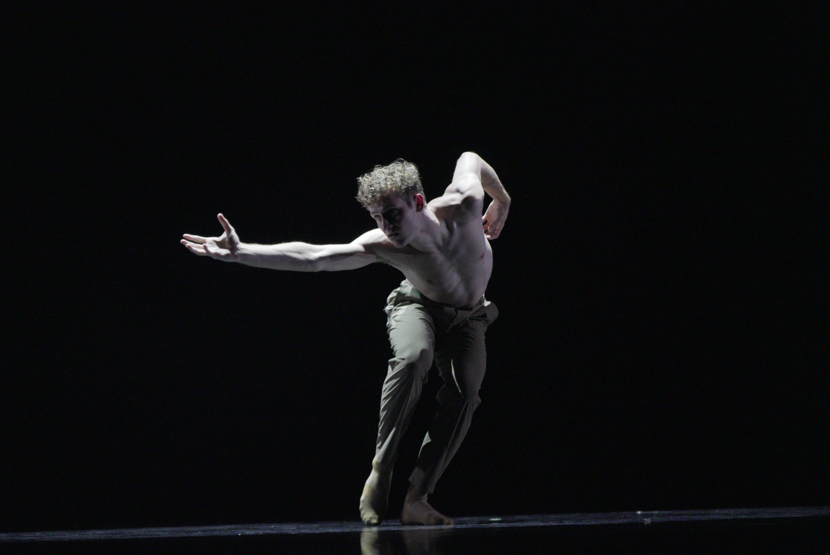 A male dancer performs on stage with a dark background, shirtless, wearing beige pants, striking a dynamic pose with one arm extended forward and the other bent behind his back.