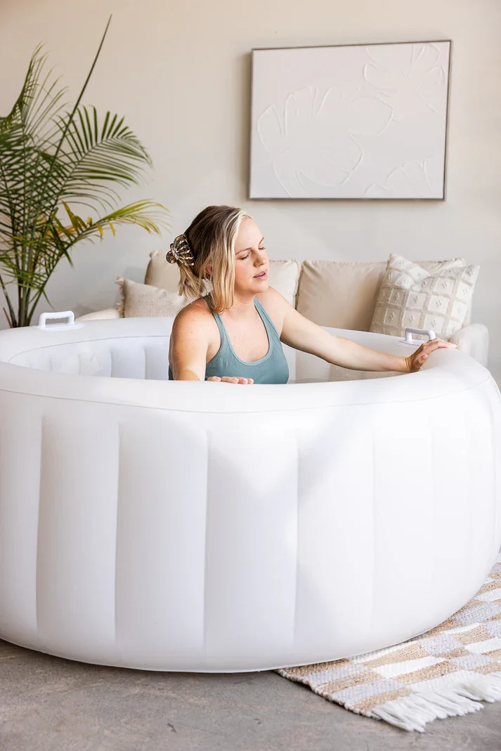 A woman soaking in a white inflatable bathtub in a living room, with a beige couch, decorative pillows, a plant, and abstract wall art in the background.