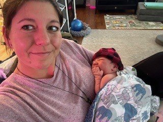 A woman (Jacquelle Amado) smiling with a newborn baby wrapped in a blanket, lying on her shoulder in a cozy indoor setting.