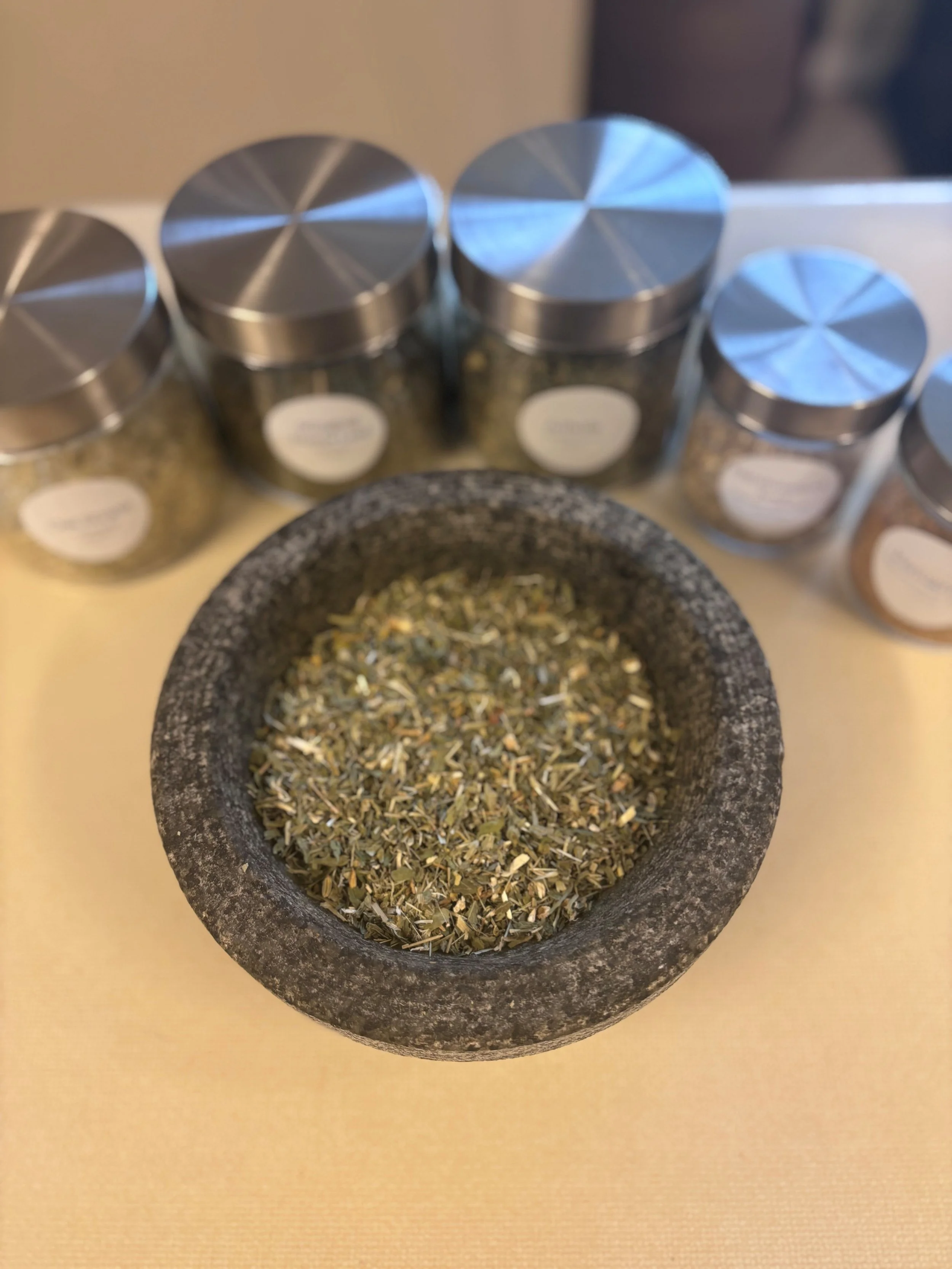 A black stone mortar filled with dried herbs in front of several glass jars with metal lids, each labeled with various herb names.