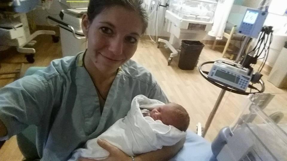 A woman (Jacquelle Amado) in a hospital gown holding a newborn baby wrapped in a white blanket, smiling in a hospital room.