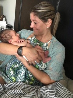 A woman (Jacquelle Amado) holding a newborn baby on her lap in a bedroom, smiling and looking at the baby, with the woman wearing a smartwatch and a floral dress.