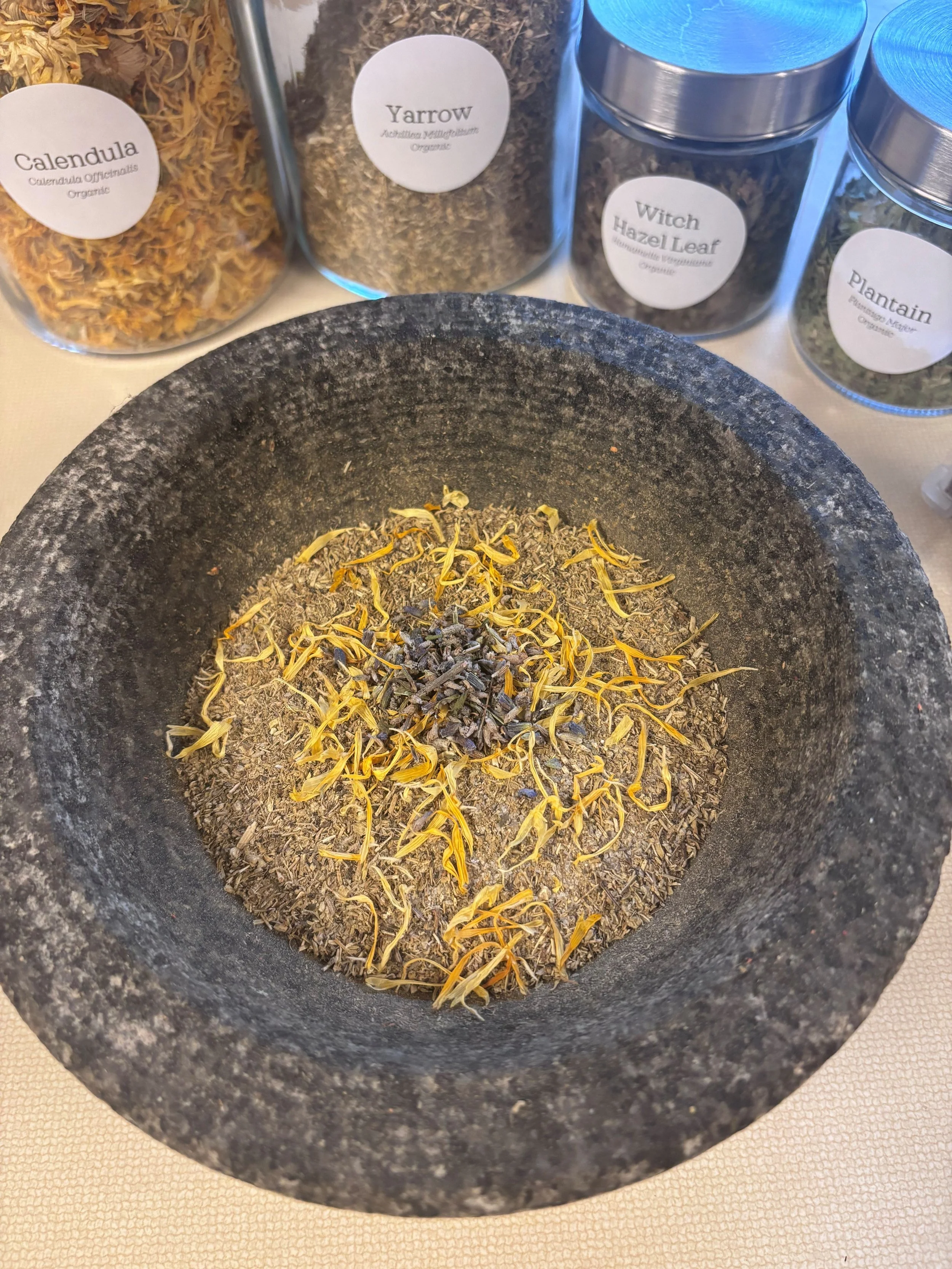 A black stone mortar with dried herbs and yellow flower petals inside. In the background, there are four labeled jars of dried herbs on a light-colored tablecloth.