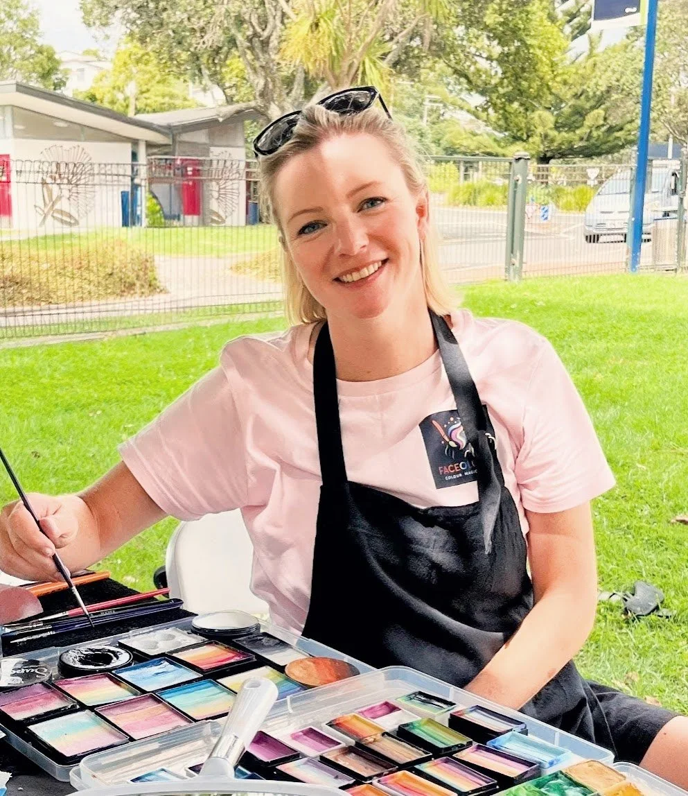A woman sitting outdoors at a table, painting with colorful watercolors. She is smiling, wearing a light pink shirt, black apron, and sunglasses on her head. Behind her is a grassy area with trees, a fence, and some buildings.