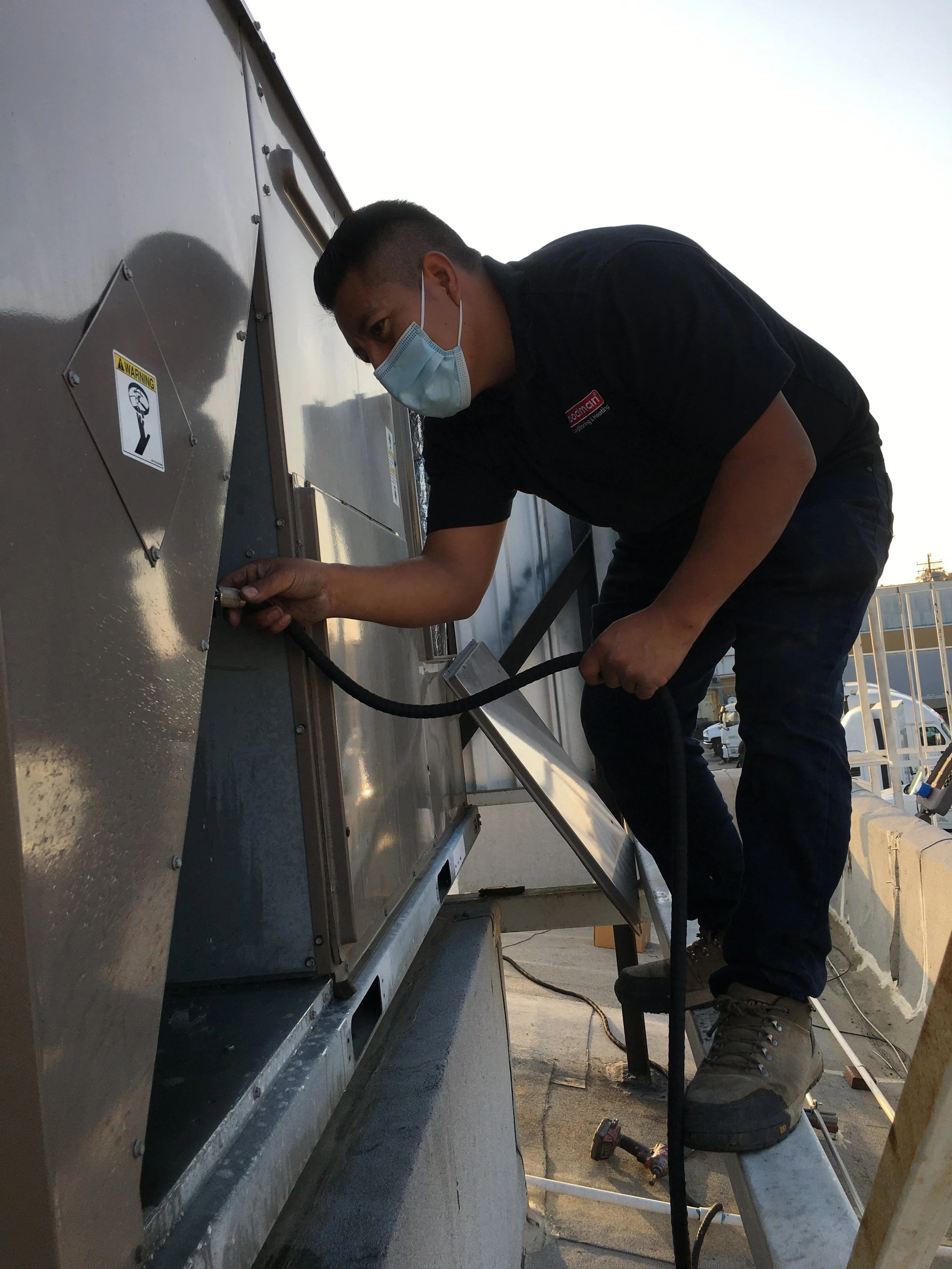 A man wearing a face mask and work boots is working on installing or repairing an industrial metal equipment outside during daylight, using a power tool.