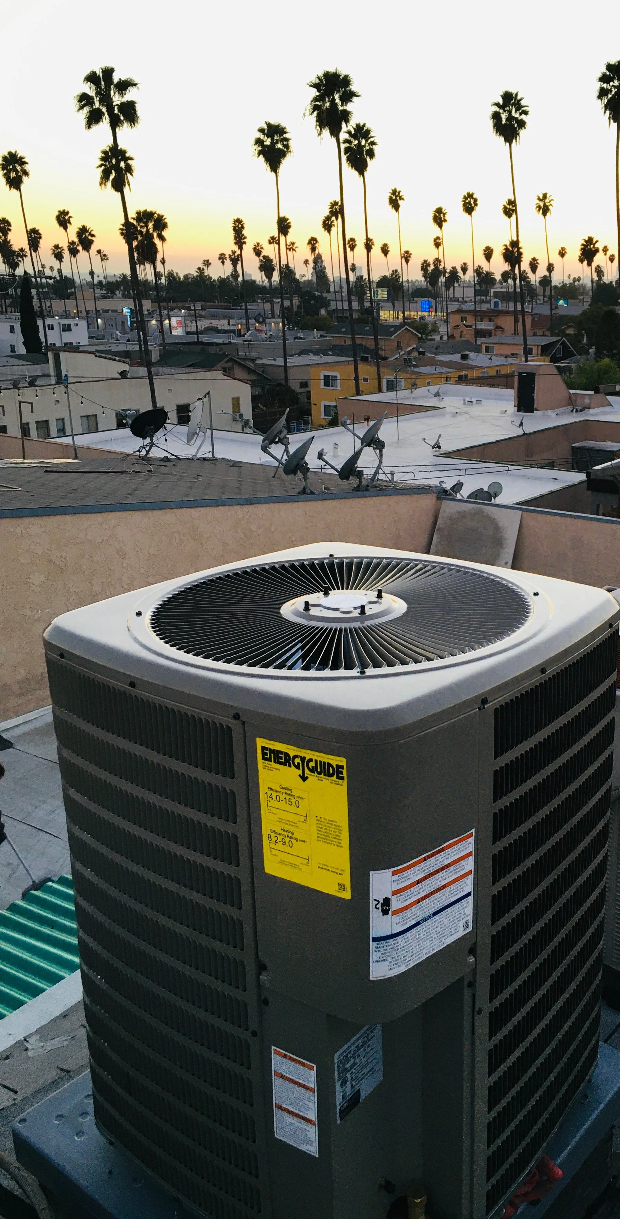 Air conditioning unit on a rooftop with palm trees and rooftops in the background during sunset.