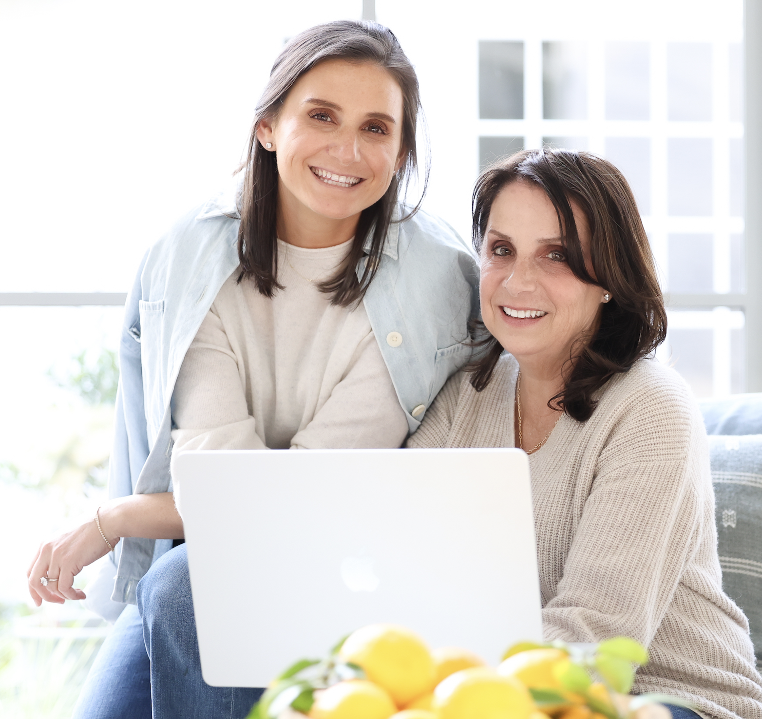 Two women with dark hair smiling at a laptop in a bright room with large windows and a bowl of lemons in the foreground.