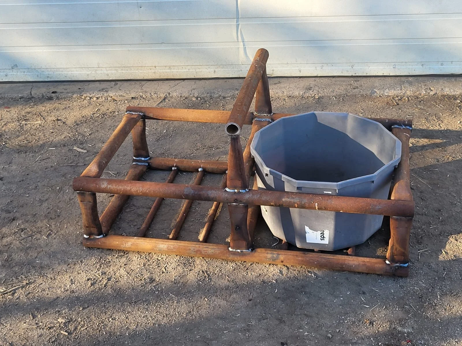 A rusted metal frame with a gray plastic container on dirt ground, in front of a light beige metal wall.