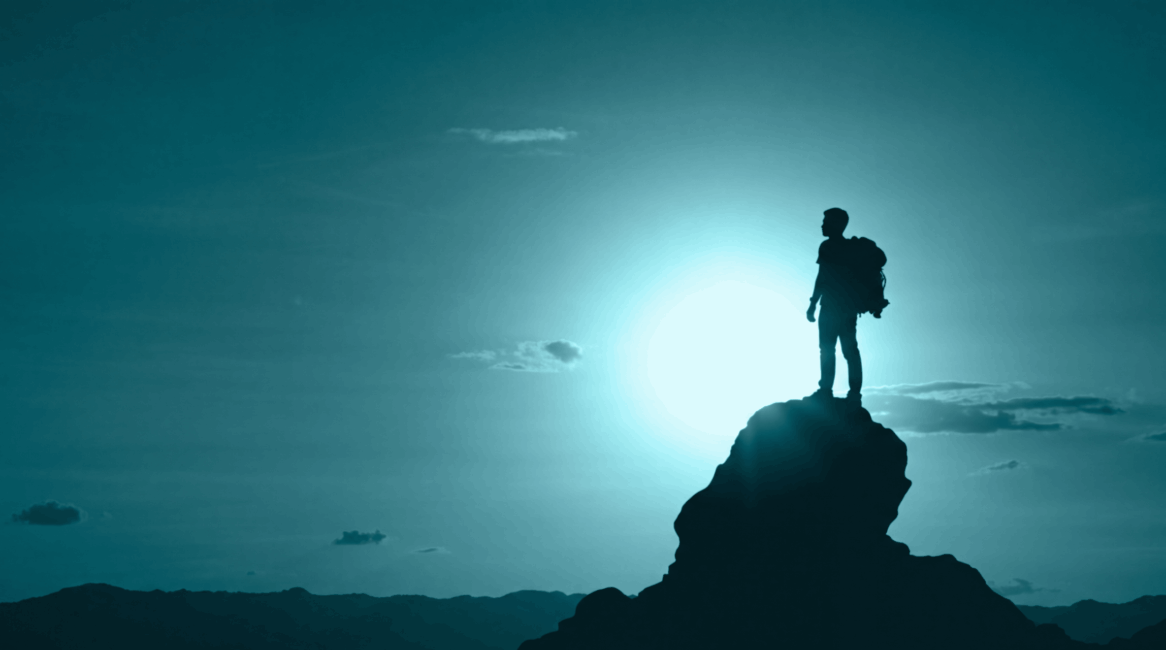 Silhouette of a person standing on a rocky hilltop against the bright sun in the sky with clouds.