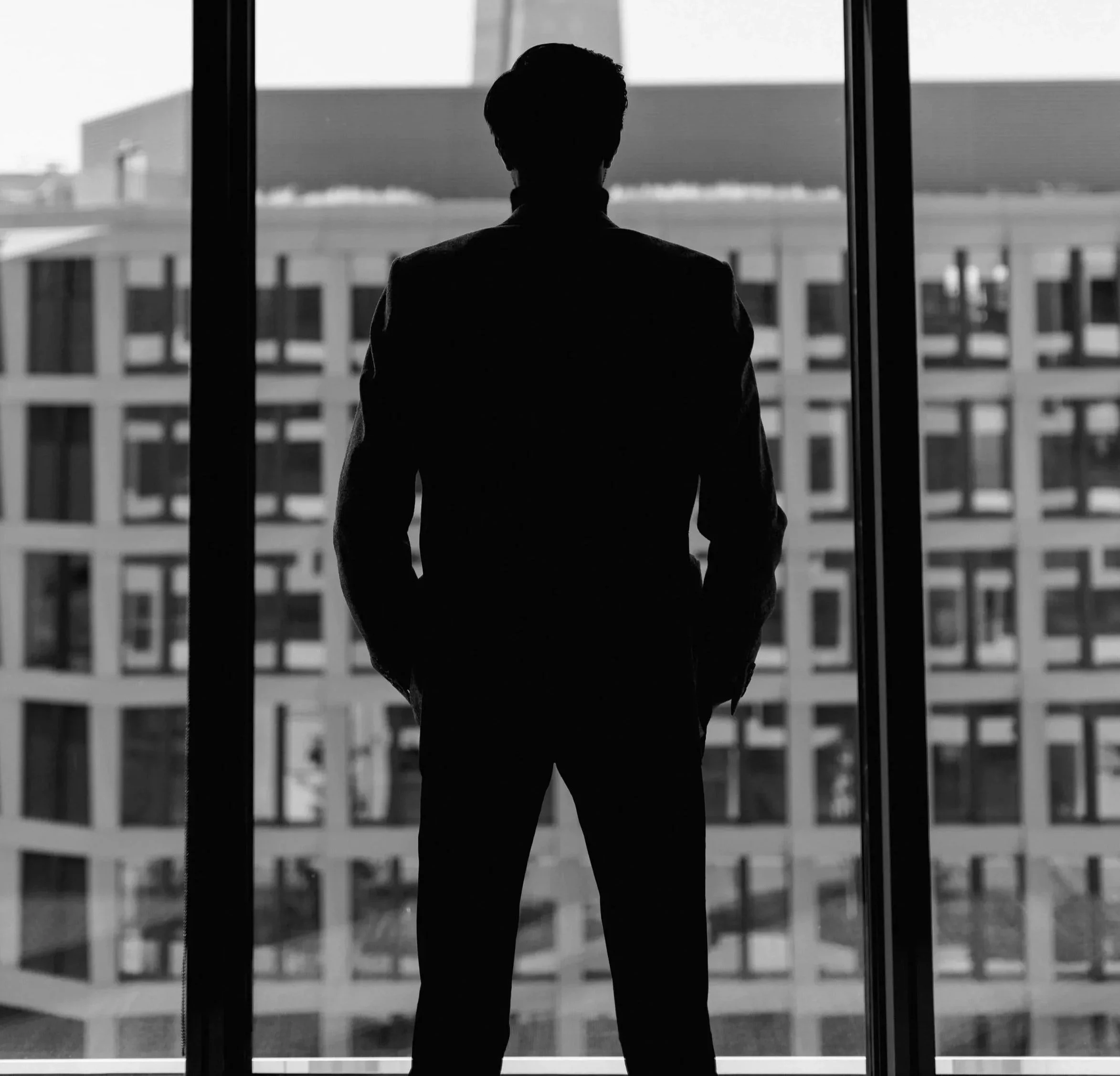 Silhouette eines Mannes in Anzug vor großen Fenstern mit Stadtgebäuden im Hintergrund.