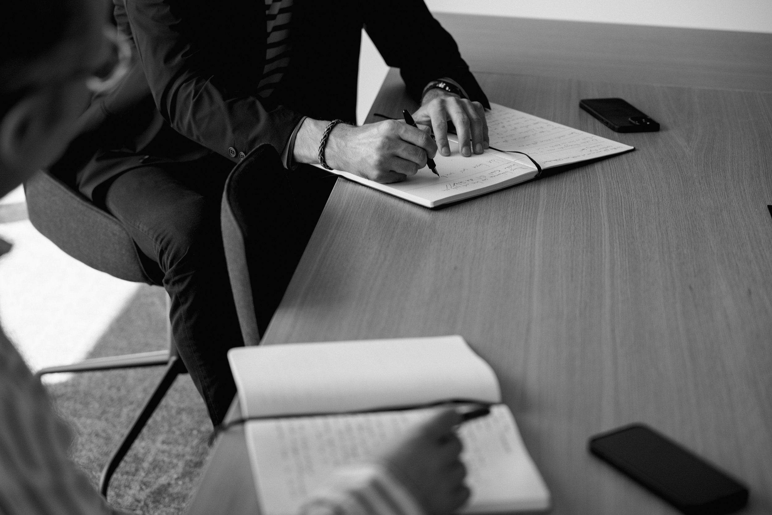 Mann schreibt in einem Notizbuch bei einer Besprechung, mehrere Mobiltelefone auf dem Tisch.