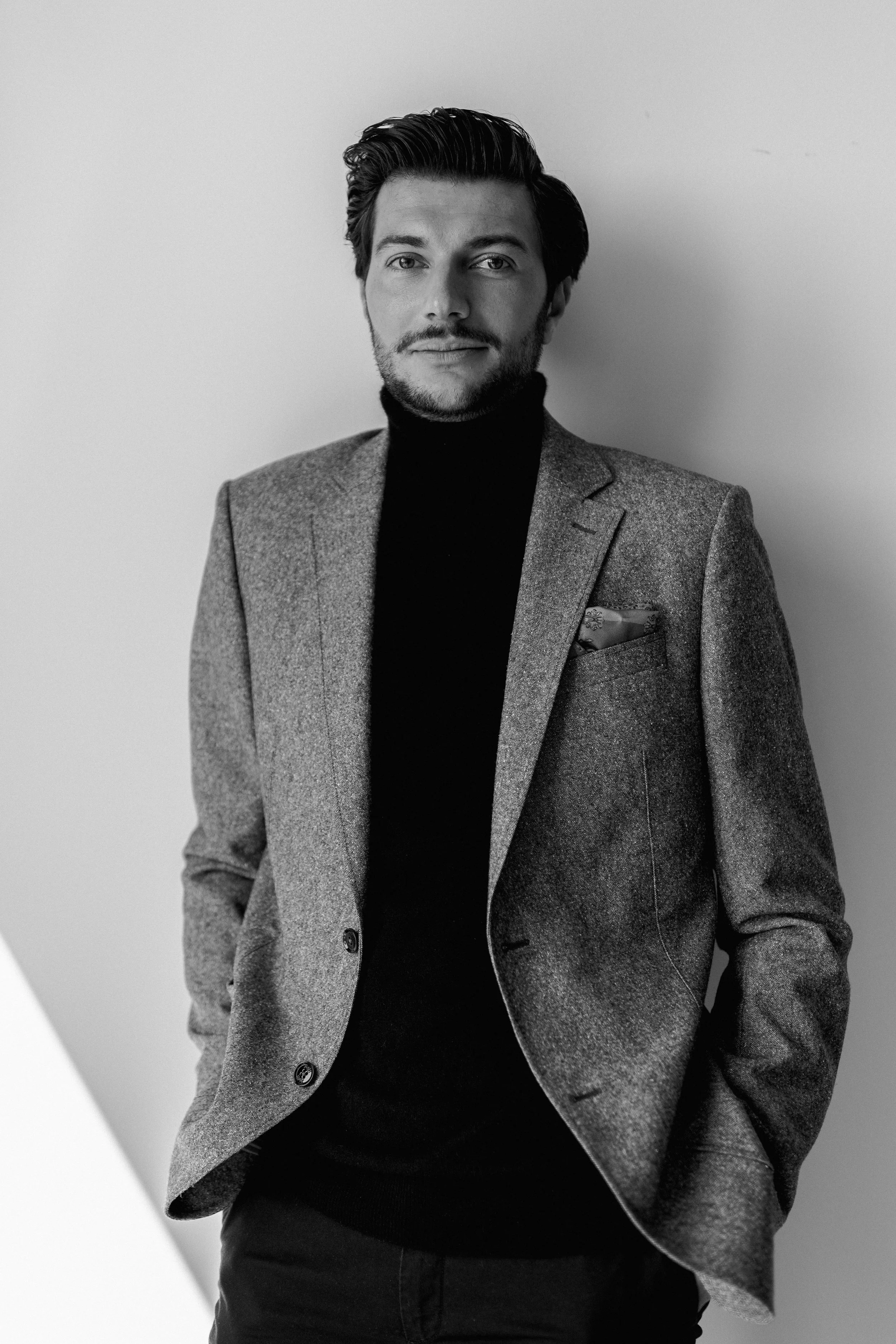 Schwarz-Weiß-Fotografie eines Mannes in einem grauen Anzug und schwarzen Rollkragenpullover, der vor einer hellen Wand steht.