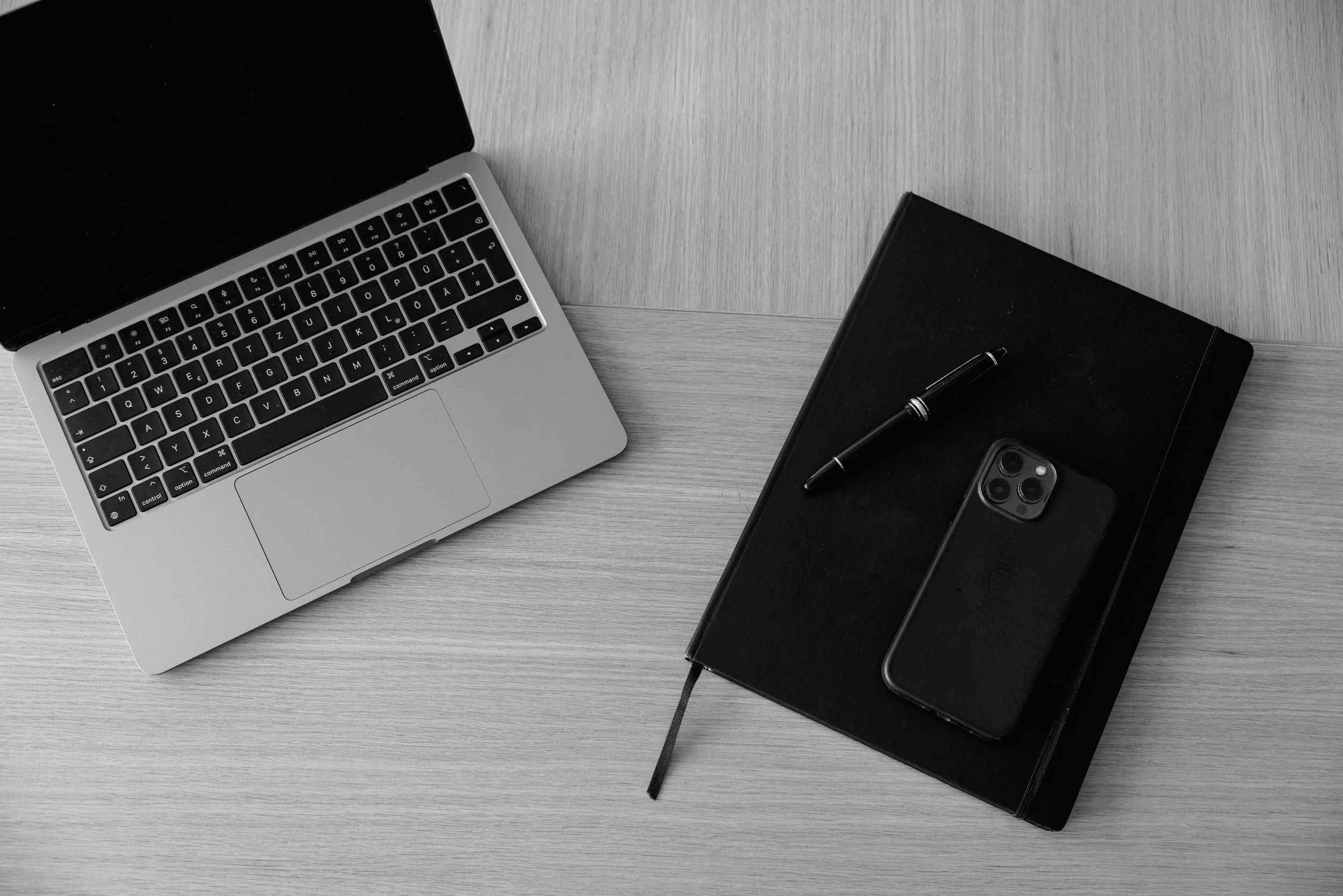 Ein Laptop, ein schwarzes Notizbuch, ein Stift und ein Smartphone auf einem grauen Holztisch.