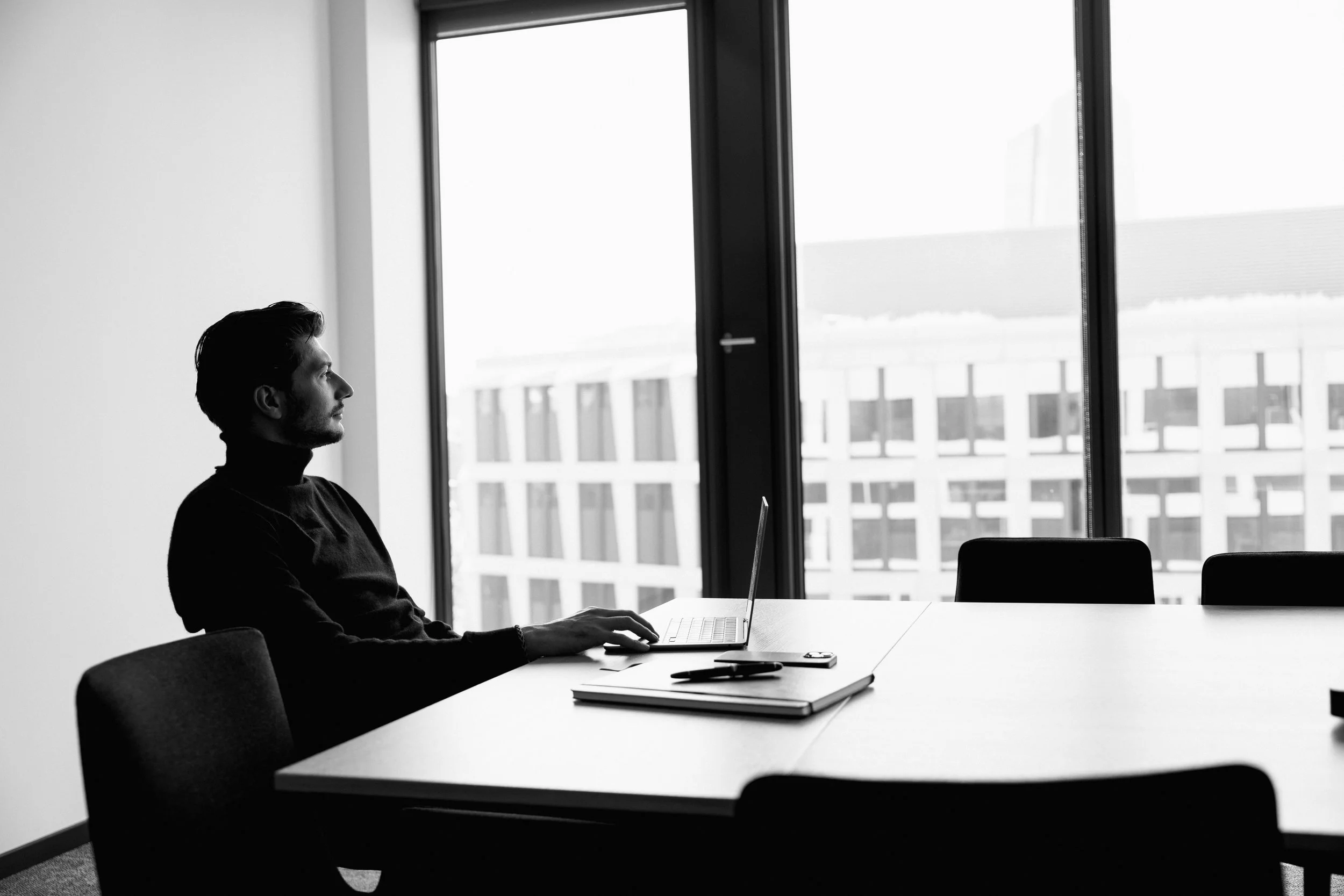 Mann sitzt alleine an einem Tisch in einem modernen Büro, schaut nach draußen durch große Fenster bei Tageslicht.