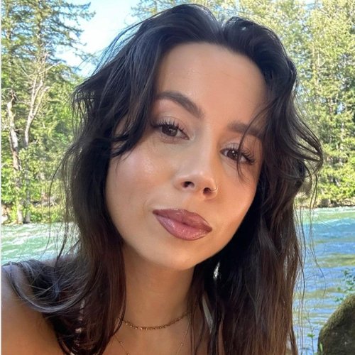 A woman with dark, wavy hair and makeup, smiling slightly, outdoors near a body of water with trees in the background.