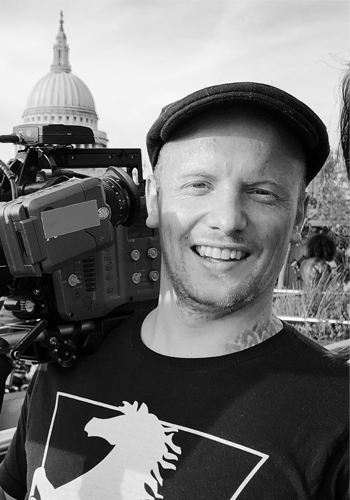 Smiling man wearing a hat and a black t-shirt with a unicorn graphic, standing outdoors with camera equipment, the United States Capitol dome visible in the background.
