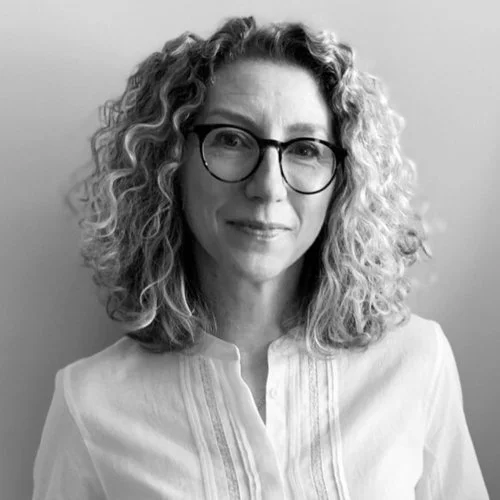 A black-and-white portrait of a woman with curly hair wearing glasses and a button-up shirt, smiling softly at the camera.