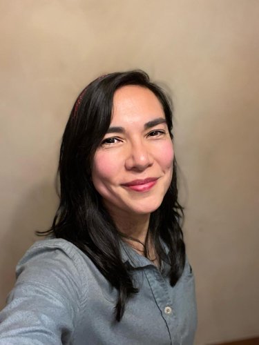 A woman with shoulder-length dark hair, smiling, wearing a gray shirt, standing against a plain beige wall.