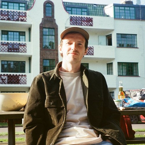 Young man sitting outdoors with modern white building in background, wearing a beige cap, white t-shirt, and dark jacket.