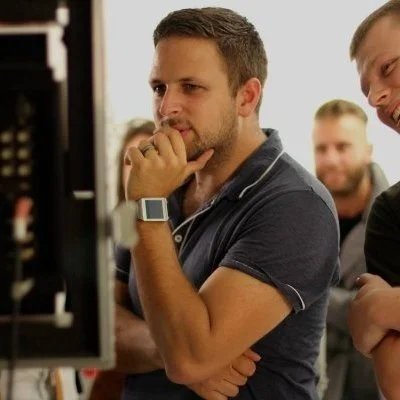 A man with short brown hair and a beard looking at a computer screen with a thoughtful expression, wearing a dark polo shirt and a smartwatch, with other men standing nearby.