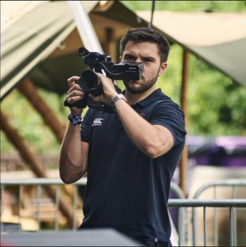A man is filming with a video camera outdoors, with a wooden structure and green trees in the background.