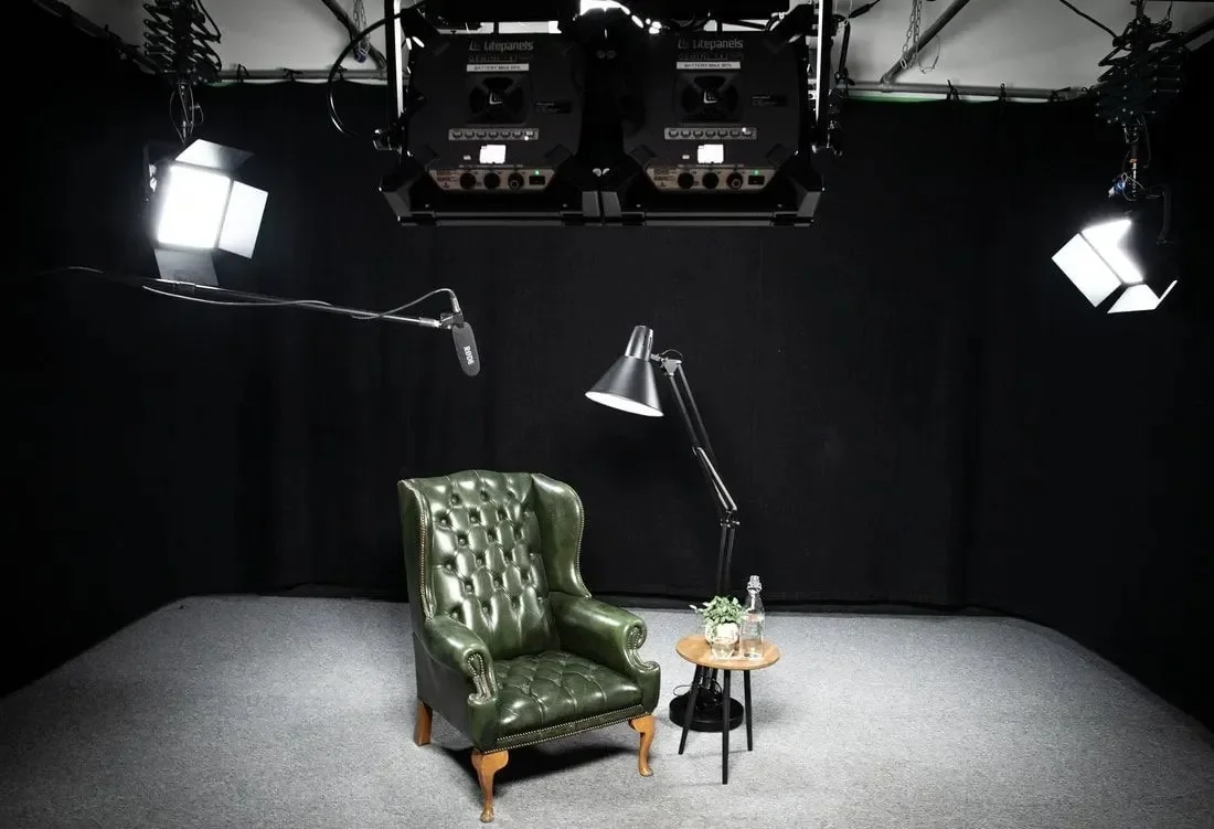 A green leather armchair with wooden legs, a small round wooden side table with a plant and water bottle, a black floor lamp, and studio lighting and equipment in a dark photography studio.