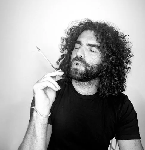 A man with curly hair and a beard holds a cigarette near his face, eyes closed, wearing a black t-shirt against a plain background.