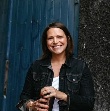 A woman with shoulder-length brown hair smiling, wearing a black denim jacket, standing outdoors against a blue fence and black wall.