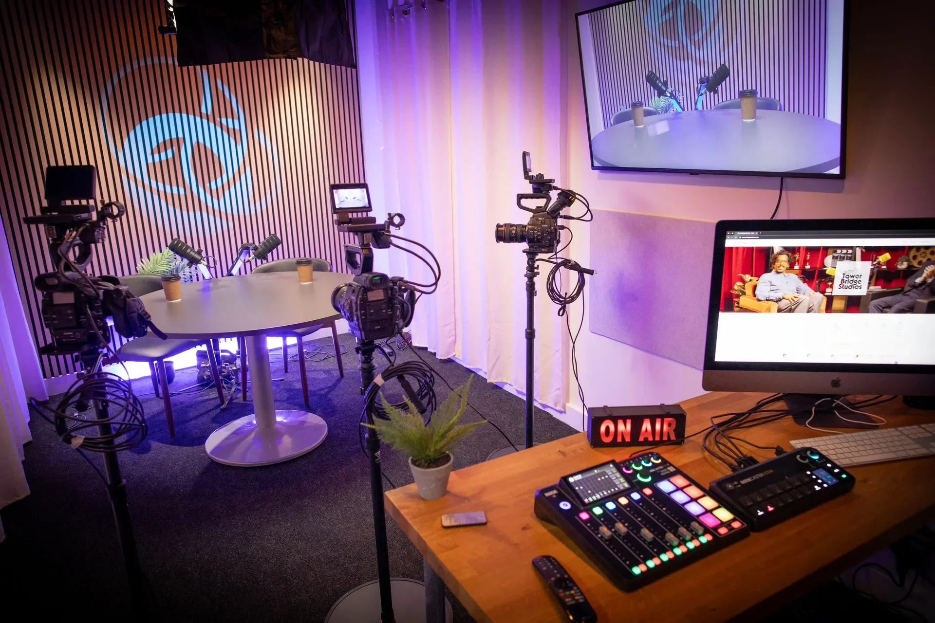 A TV screen showing a podcast setup with two microphones and coffee cups on the table, a filming camera, a computer monitor displaying a podcast page, and an 'ON AIR' sign in a recording studio.