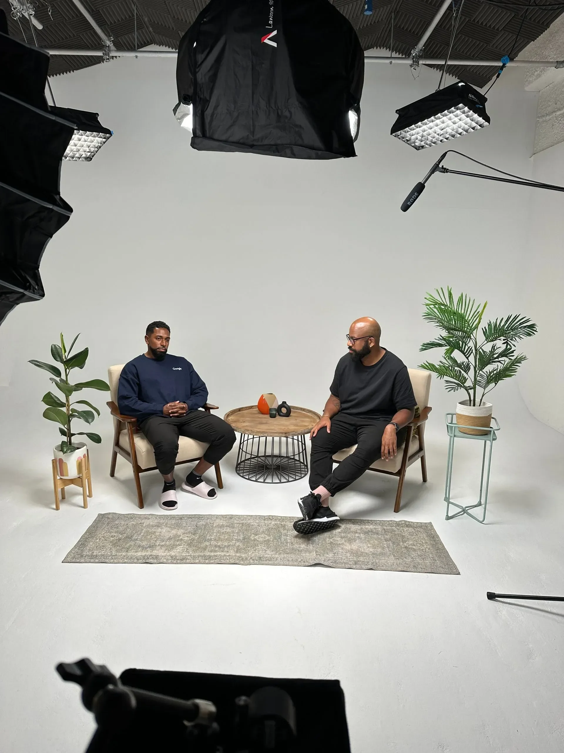Two men sitting on beige chairs with wooden frames, having a conversation in a studio setting. Between them, there is a round wooden table with a kettle and a black kettlebell on it. There are two potted plants, one on each side of the setup. Overhead studio lights and a microphone on a boom arm hang from the ceiling, and a camera is positioned in front of them.
