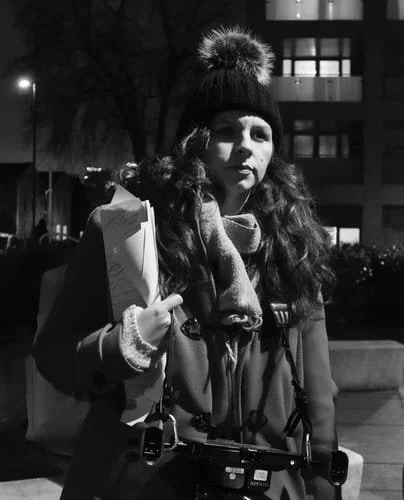 A young woman wearing a winter hat with a pom-pom, a scarf, and a coat, standing outdoors at night with buildings and streetlights in the background. She has a camera hanging around her neck and is holding some papers or a notebook.