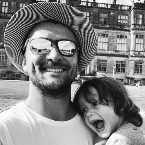 A man with a hat and sunglasses smiling for a selfie with a young girl who is laughing and leaning against him, in front of a historic building.