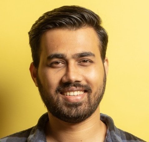 Smiling man with dark hair and beard against yellow background.