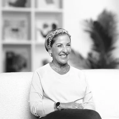 A woman smiling and sitting on a couch in a living room with shelves and plants in the background.