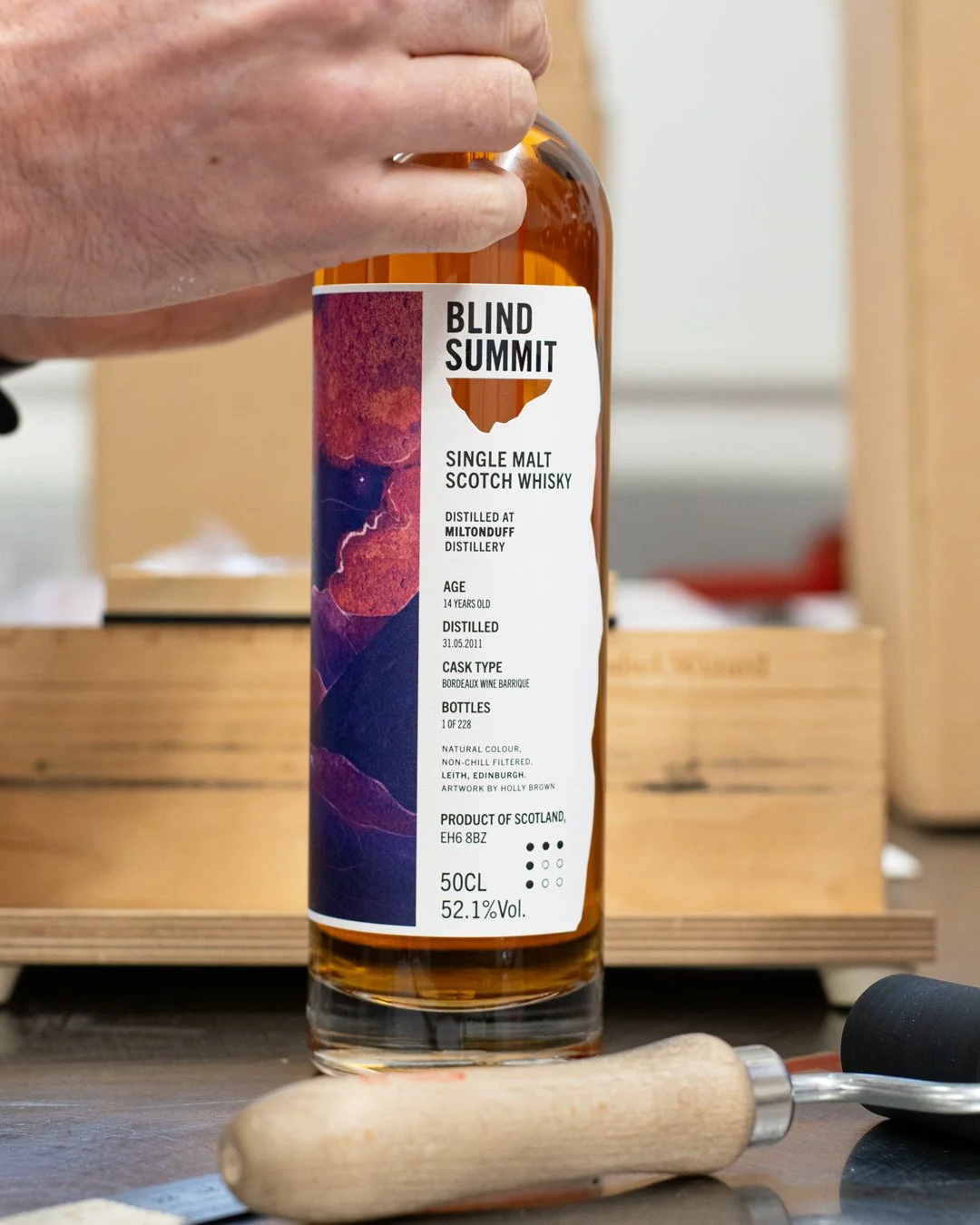 A person holding a bottle of Blind Summit single malt Scotch whisky. The bottle label provides details including age, distillation date, cask type, bottle number, alcohol percentage, and artwork by Holly Brown. The scene includes a wooden box, a cork