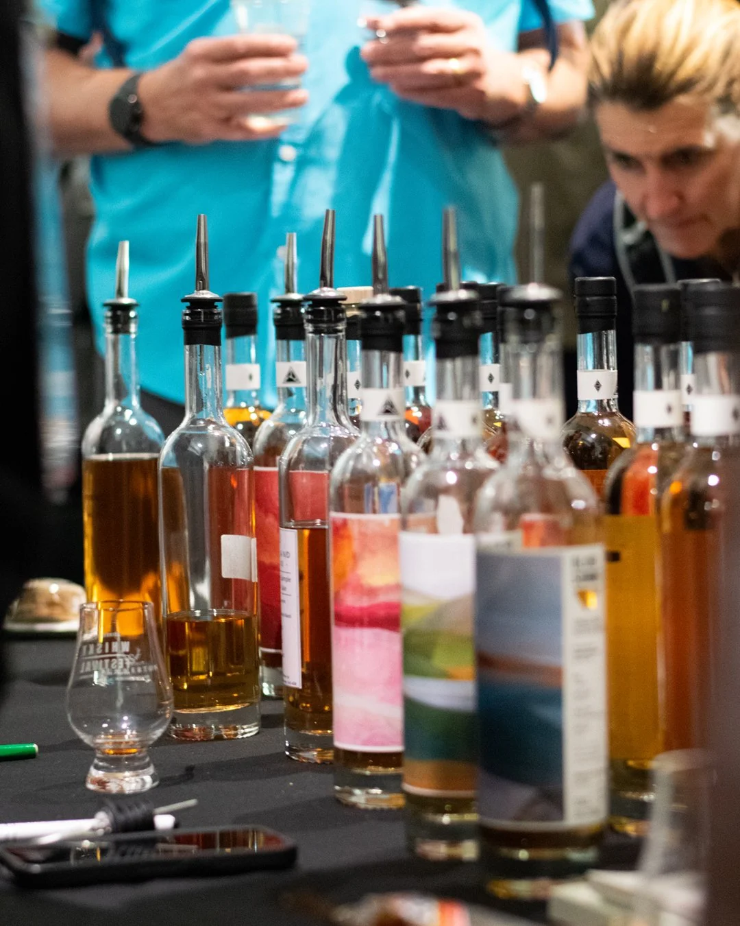 Multiple bottles of flavored whiskey with pipettes inserted on a table, with two people in the background examining the bottles.