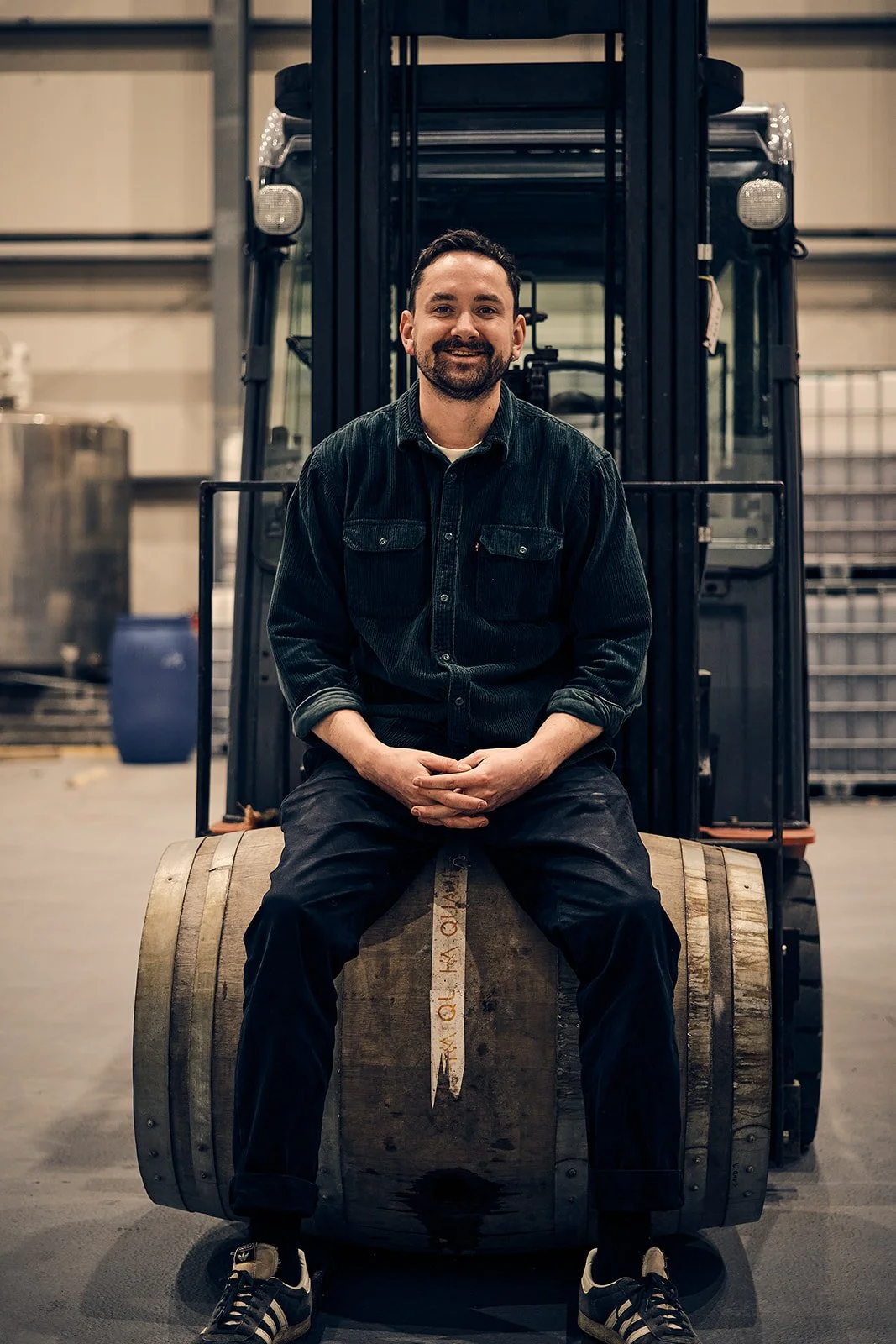 A man sitting on a wooden barrel in an industrial warehouse, with a forklift behind him.