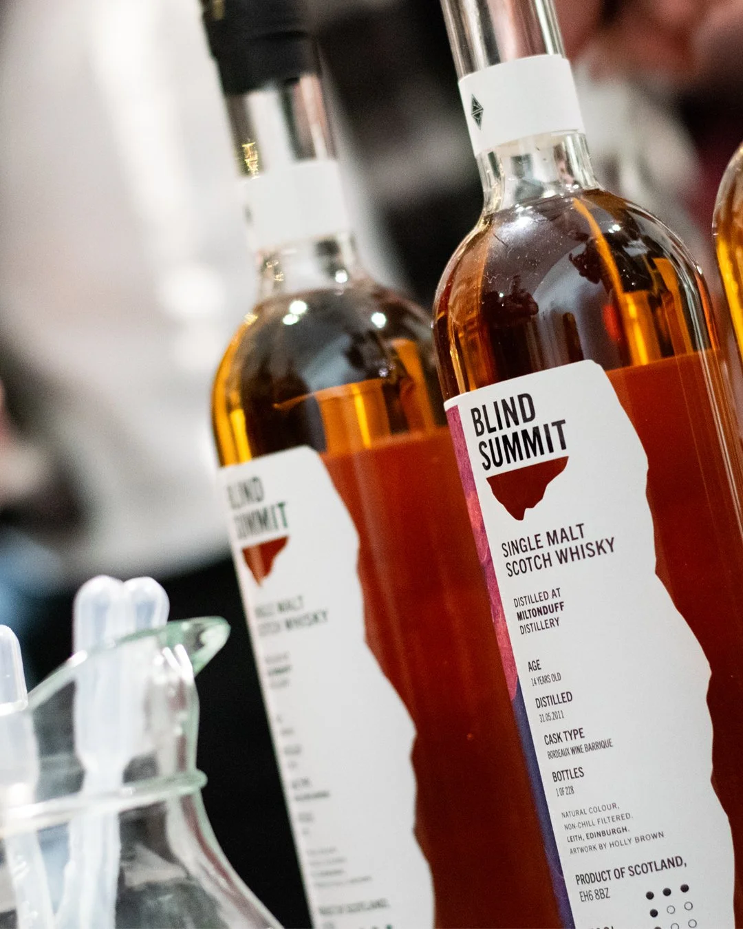 Close-up of two bottles of Blind Summit single malt Scotch whisky on a bar counter with a blurred background.