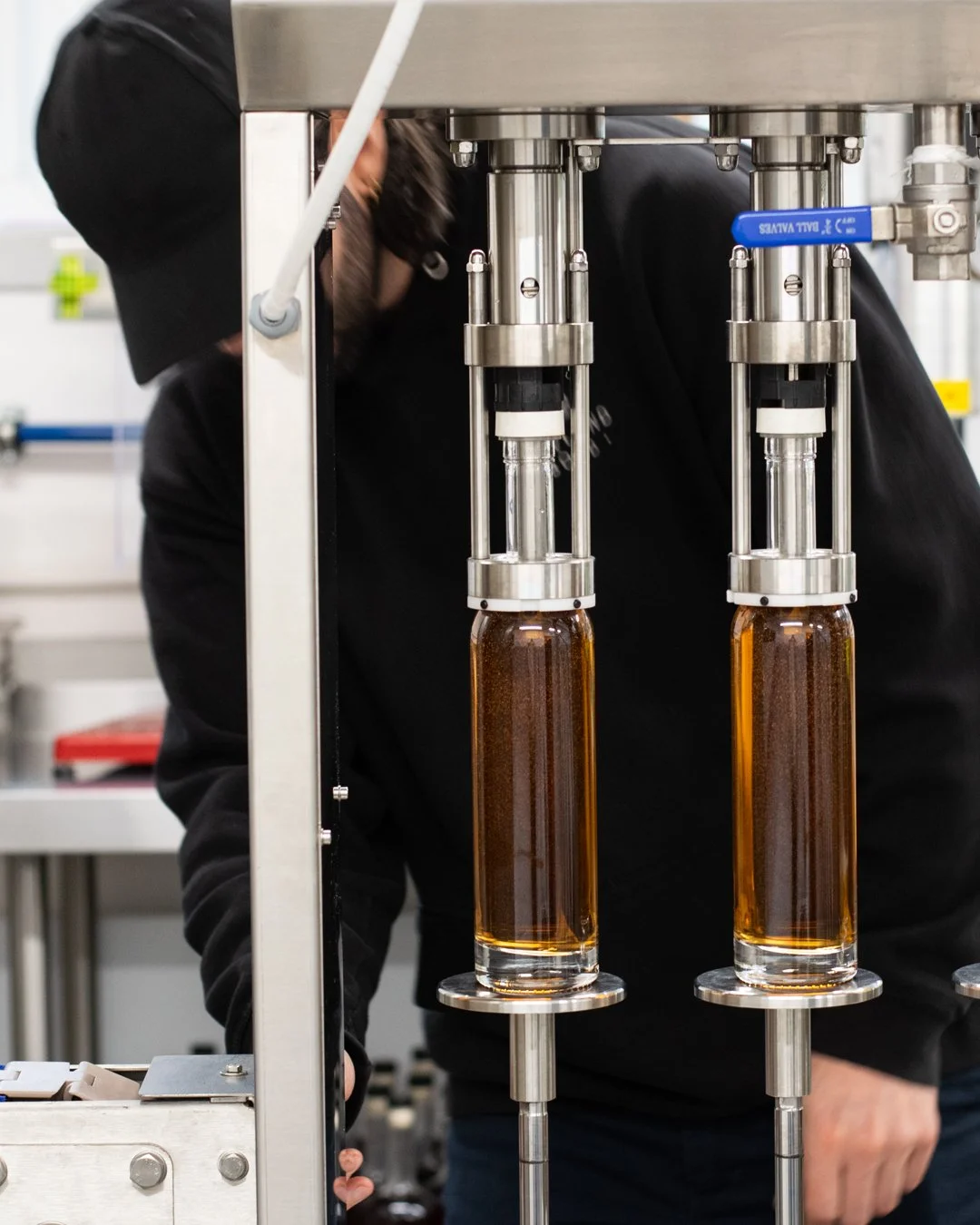 Industrial setting with two vertical glass bottles filled with amber liquid, using machinery for filling or dispensing, with a person partially visible in the background.