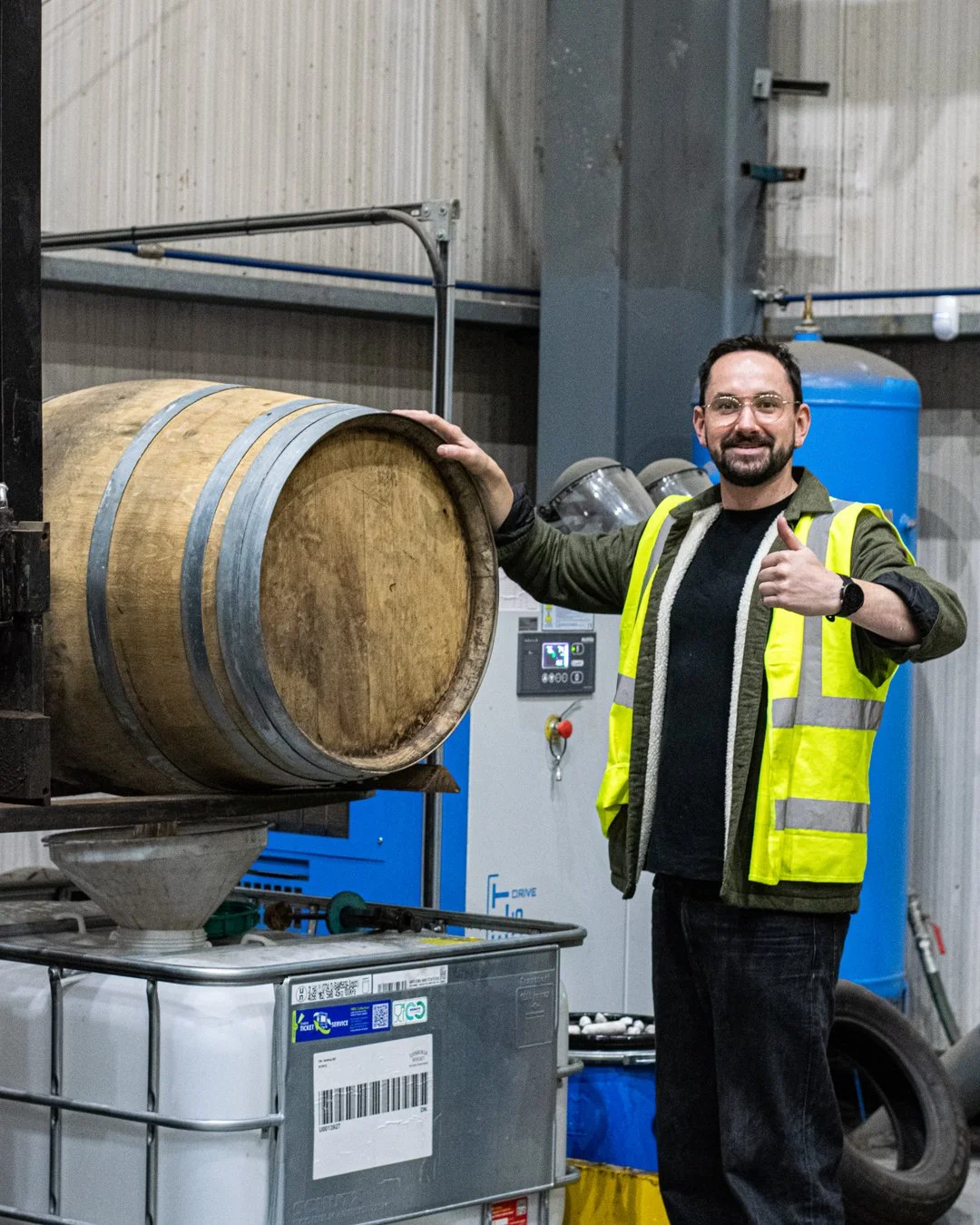 A man in safety vest giving a thumbs up and leaning on a barrel in an industrial setting.