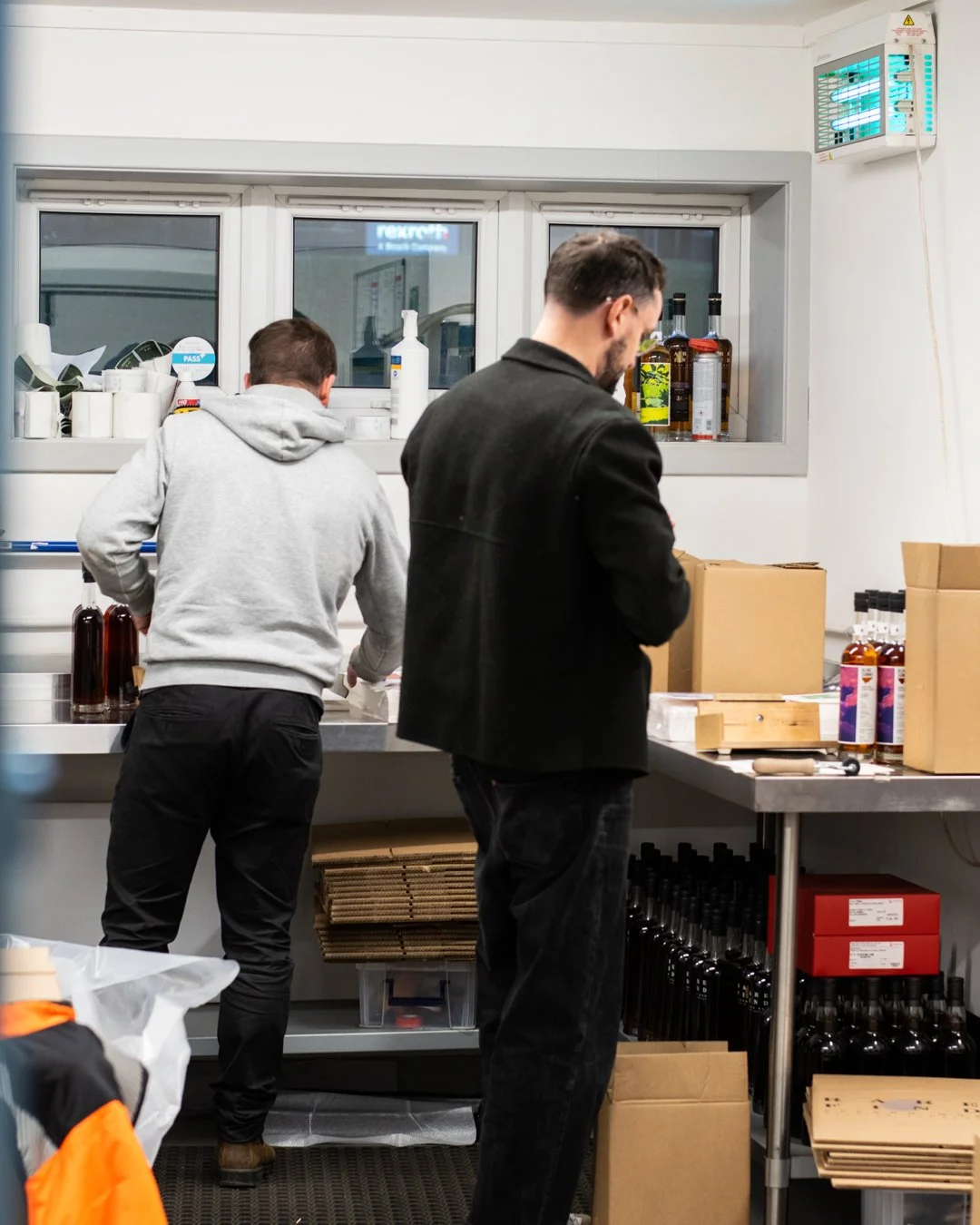Two men working behind a counter in a kitchen or bar area, with bottles, boxes, and equipment on the counter and shelves.