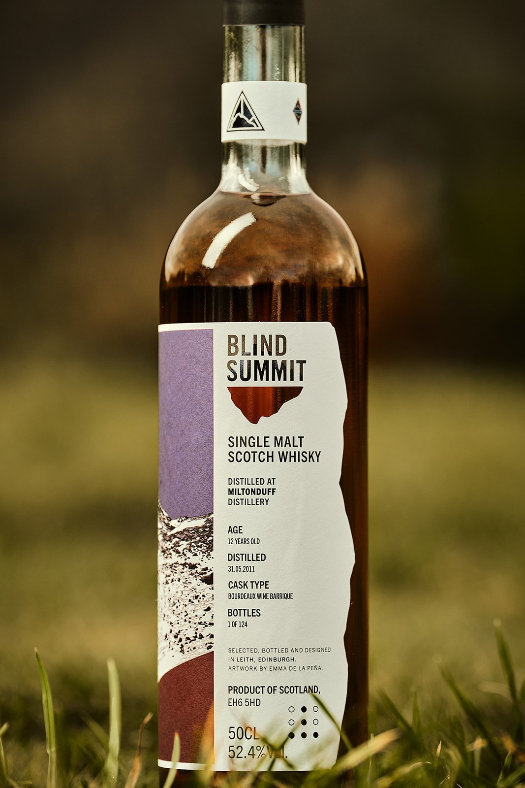 Bottle of Blind Summit single malt Scotch whisky with a white label featuring purple, brown, and black details, placed outdoors on grass.